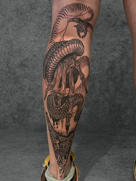 A tattoo showing an intricate skeletal dragon design wrapping around the calf of a male, created in detailed black ink style