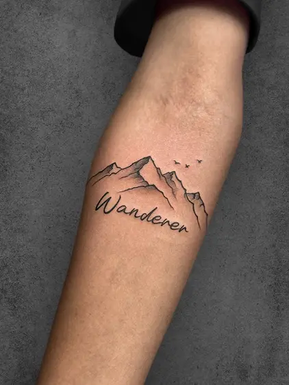 A minimalistic tattoo of a mountain range with the word 'Wanderer' beneath it, and small birds flying above, done in fine line art, placed on the forearm