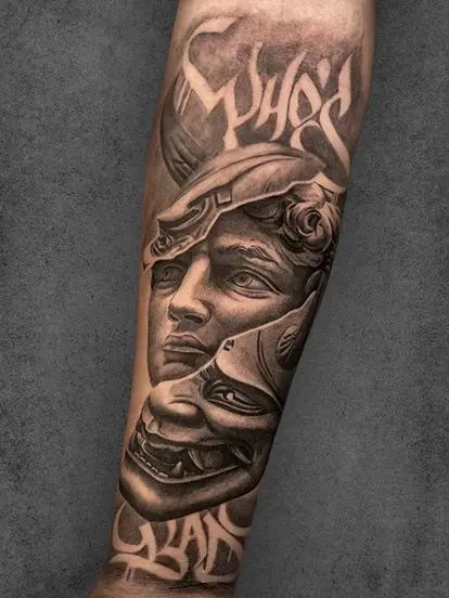 A striking arm tattoo combining a human face with a mythological mask and layered text, exploring themes of identity and duality
