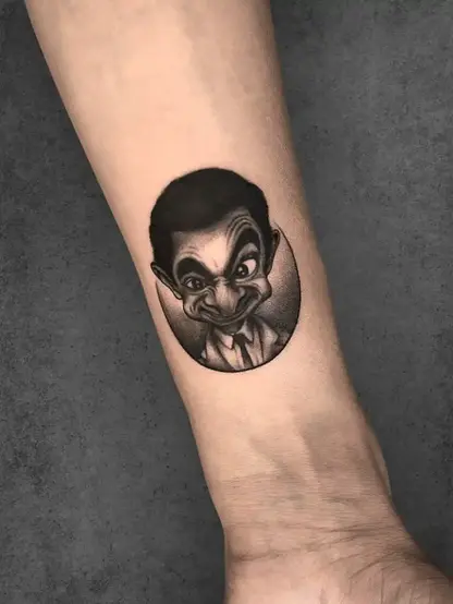 Caricature-style Mr. Bean tattoo with exaggerated expression, black and grey ink on forearm