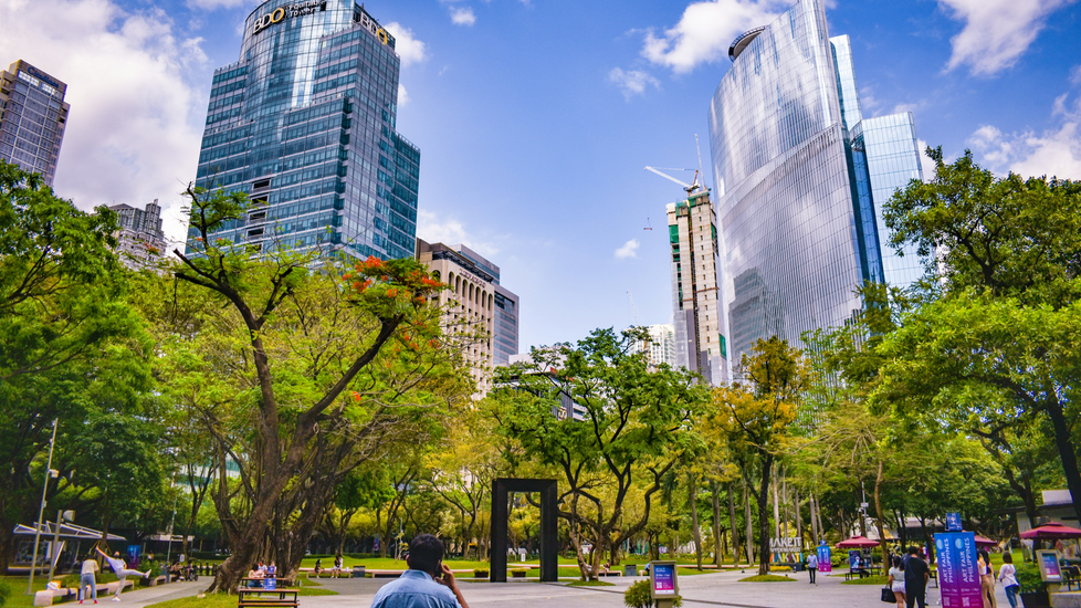 City park with trees and a walking path surrounded by tall modern skyscrapers under a bright blue sky