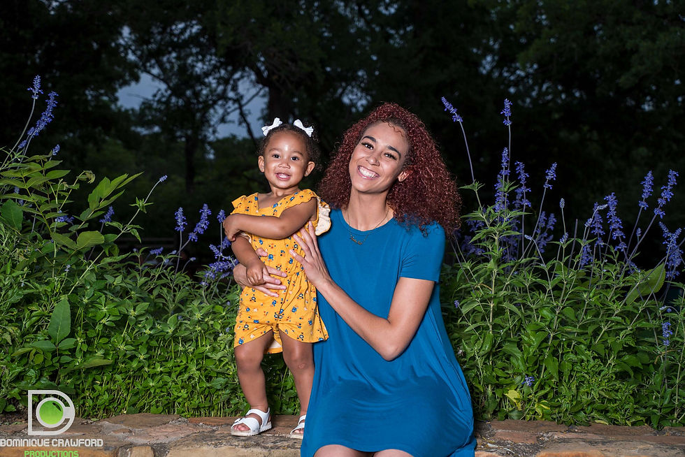 Woman in a blue dress smiling with a toddler in a yellow floral outfit outdoors among purple flowering plants at dusk