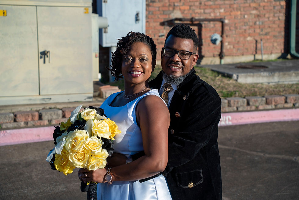 Smiling bride in a white dress holding a bouquet, embraced by the groom in a black and gold suit, posing outdoors