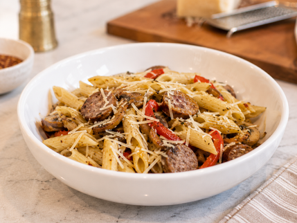 Bowl of penne pasta with sausage, red peppers, and grated cheese on a countertop, with grater and cutting board in background