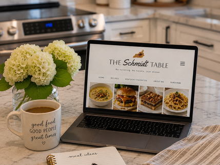 Cozy kitchen counter with laptop open to The Schmidt Table, coffee mug, hydrangeas, and meal ideas notebook.