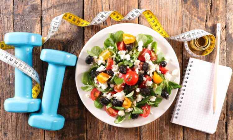 Plato de ensalada, mancuernas, cinta métrica y block de notas sobre una mesa de madera