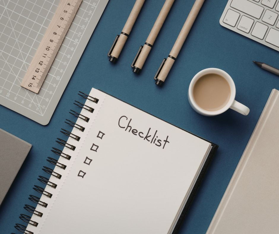 Notebook labeled "Checklist," pens, keyboard, cup of coffee on a blue surface. Ruler and cutting mat add an organized, productive mood.