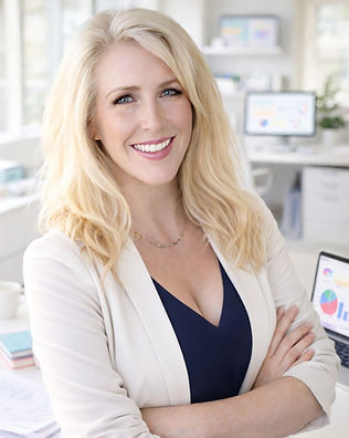 Julie Fisher, Founder and Fractional CMO of Fisher Marketing Services smiling in a white blazer and navy blouse with a gold necklace. Her shoulder length blonde hair slightly curled at the ends. She is inside an office with a window and a couple of white office desks with computer monitors behind her .
