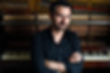 Emiliano Messiez - sitting in front of a piano with his arms crossed.