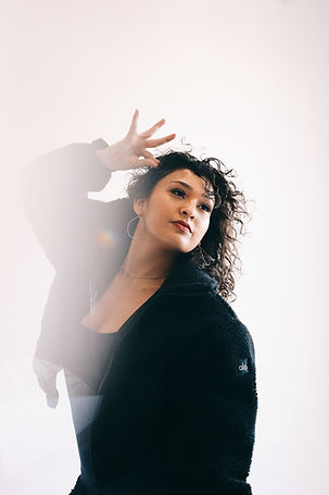 Female presenting person of latin descent, with long brown curly hair with gold hoops and makeup wearing a black furry coat. In a dance position curving her spine to the left, splaying her right hand in a teacup position.