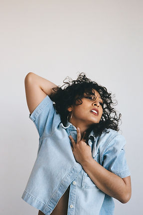 Female presenting person of latin descent, with long brown curly hair, with gold hoop earrings wearing a denim shirt. In a dance position with her head to the right side, right hand on clavicle area left hand behind her head.