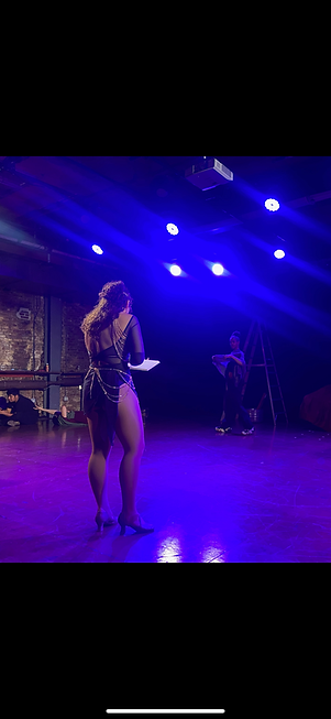 Alyssa (a female presenting person of latin descent with long brown curly hair.) with her back facing the camera wearing a blue leotard and skirt with gold garments holding a note book in blue lighting.