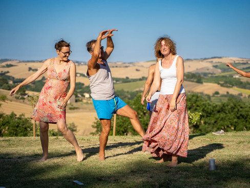 Two women and a man in summer clothes move joyfully and exuberantly to a dynamic meditation, outside in a meadow, with a beautiful view of the Italian hills.