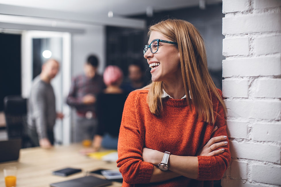 A woman looking to the side and smiling in the office, with colleagues standing in the background