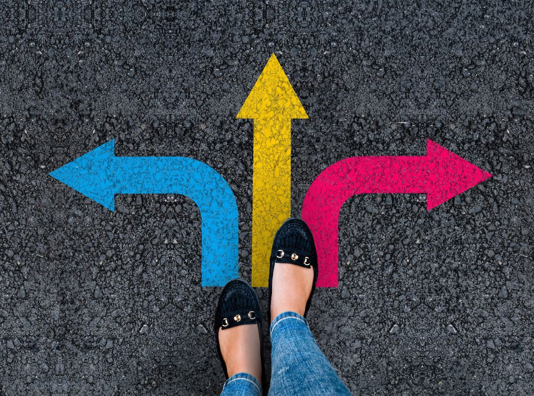 Person standing at a crossroads with arrows pointing in different directions, representing different paths toward a goal.
