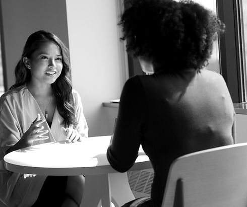 Two women sit across from each other at a table in a bright, window-lit space, engaged in conversation.