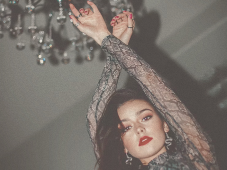 Woman confidently posing beneath a crystal chandelier wearing an elegant patterned dress and bold lipstick.