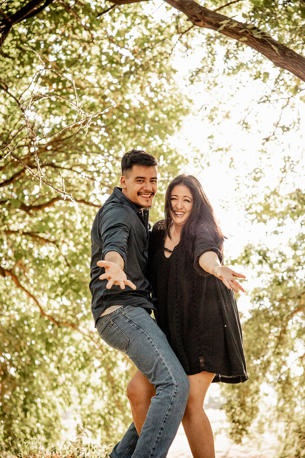 Smiling young couple under a sunlit tree, arms outstretched toward the camera in a playful outdoor pose.