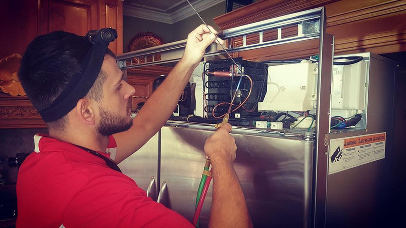 A AA Appliance Repair technician working on a component on top of the refrigerator.