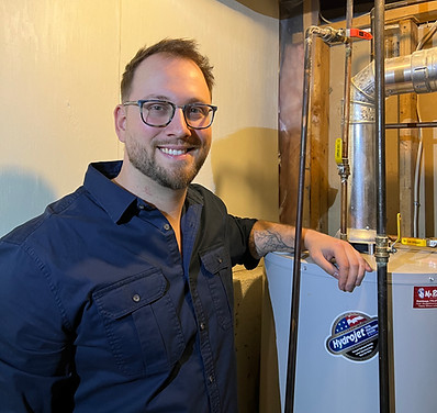 A man in a blue shirt, Jonah, stands with his arm on a hot water tank