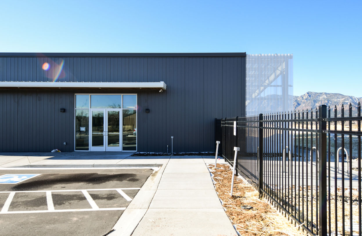 Front entrance of a dark metal building with glass doors, sidewalk access, and black metal fencing.