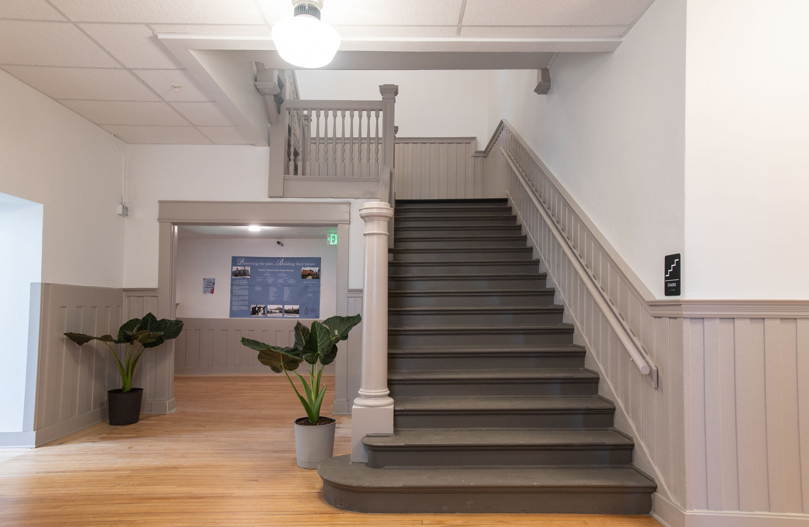  Gray-painted wooden staircase with matching wainscoting, light wood floors, and potted plants in a renovated historic building.