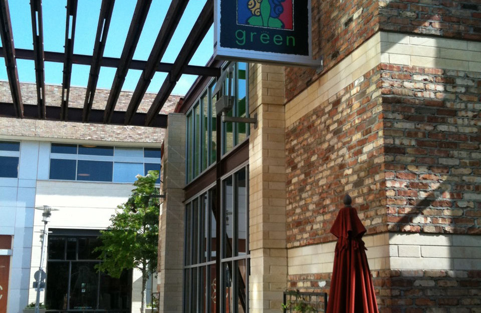 Outdoor café seating under a wooden pergola with masonry columns, storefront signage, and tables along a pedestrian walkway.