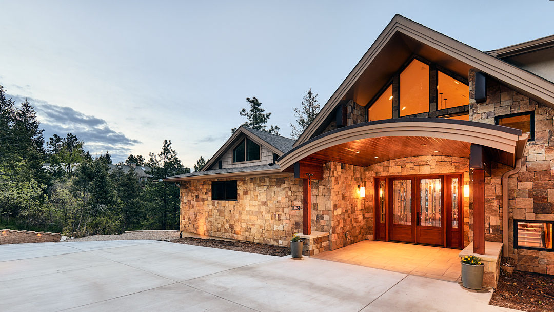 Front entry of a custom home at dusk featuring stone walls, wood beams, and warm exterior lighting.