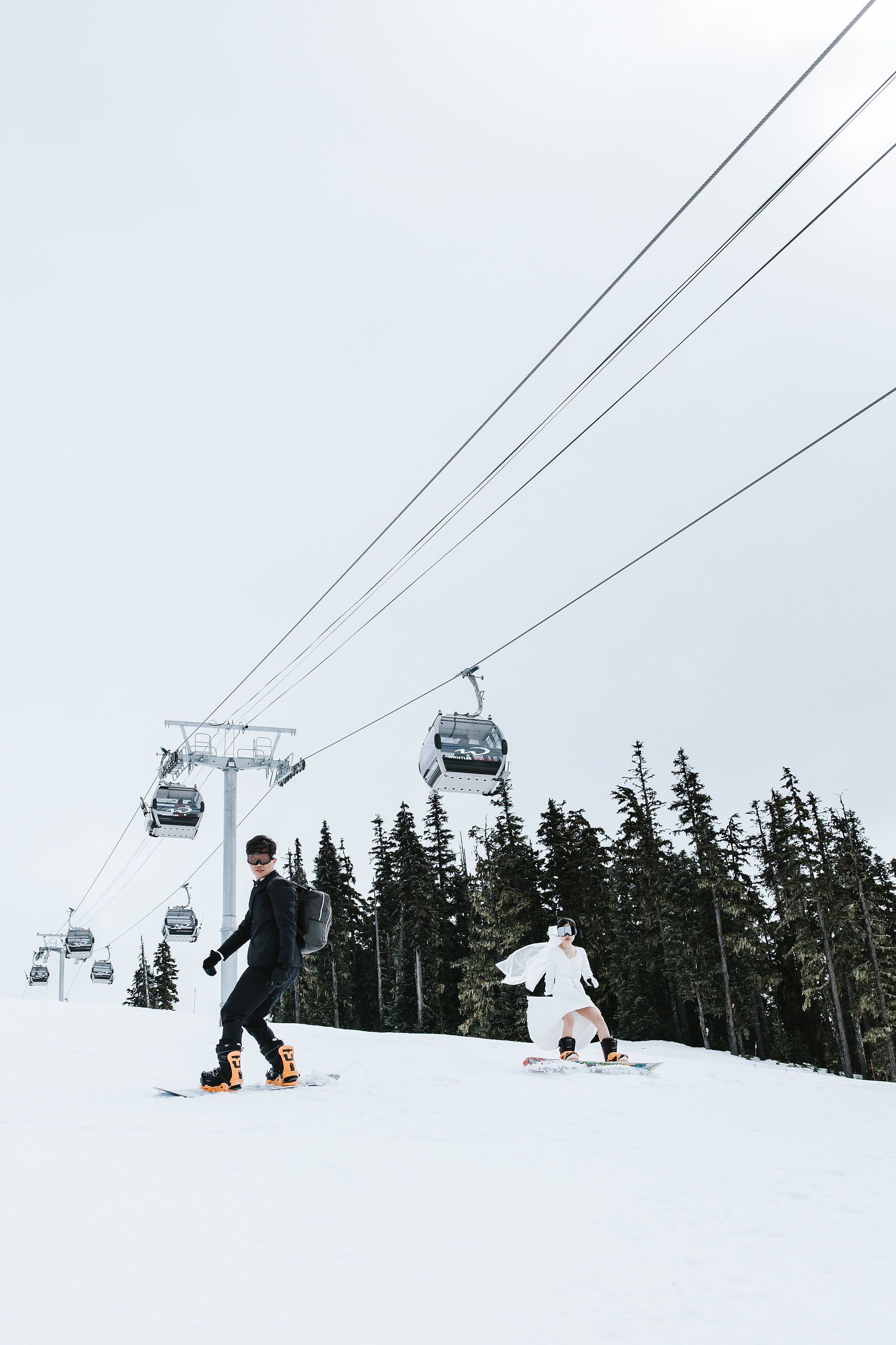 Engaged couple snowboarding down the slopes with the bride’s wedding dress flowing in the wind at Whistler Blackcomb — breathtaking and adventurous winter engagement moment.