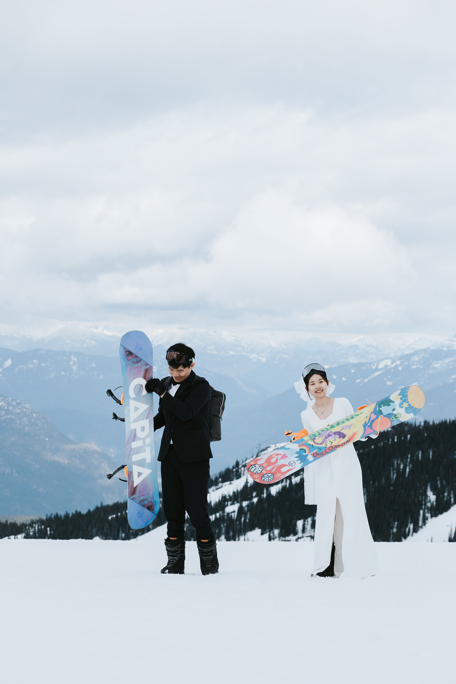  Engaged couple holding up their snowboards with the snow-covered mountains behind them at Whistler Blackcomb — adventurous and triumphant snowboard engagement moment.​​​​​​​​​​​​​​​​