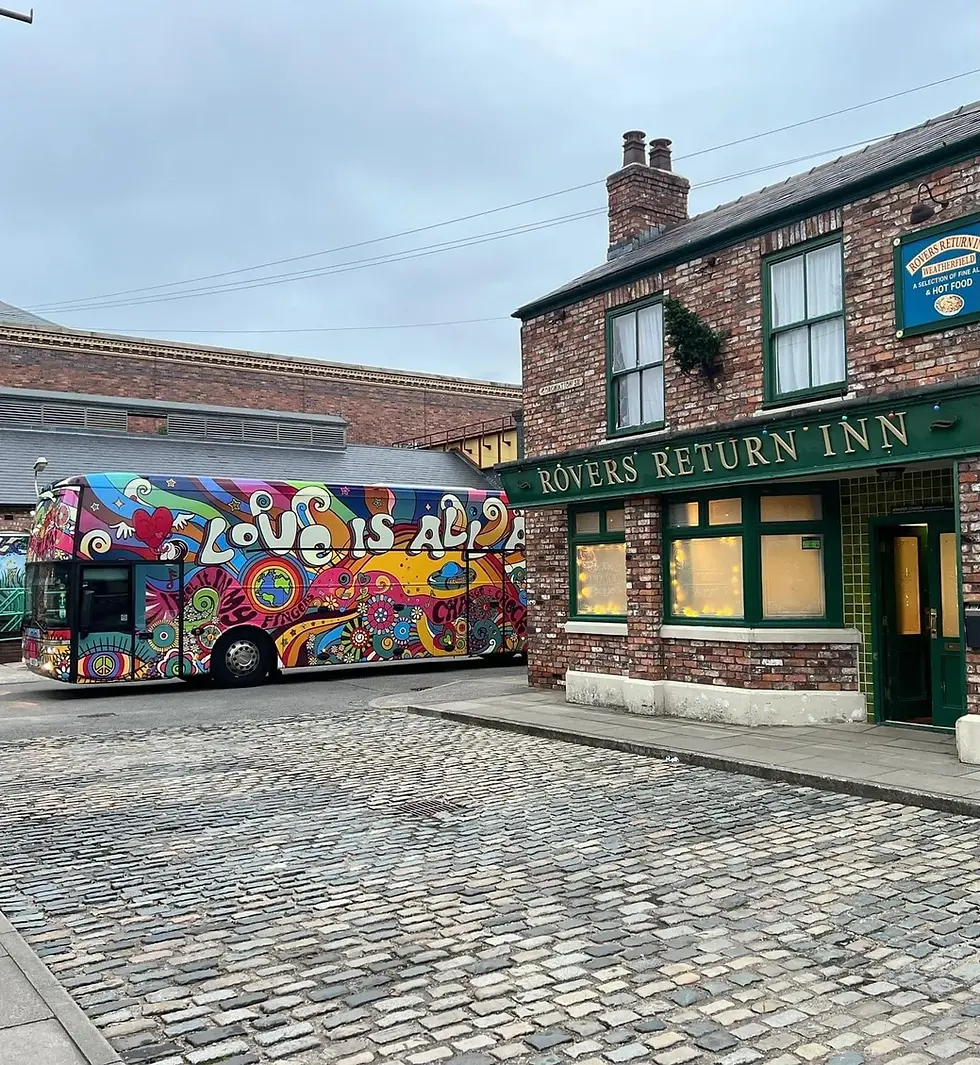 Colorful bus 'LOVE IS ALL' outside iconic 'ROVERS RETURN INN'.