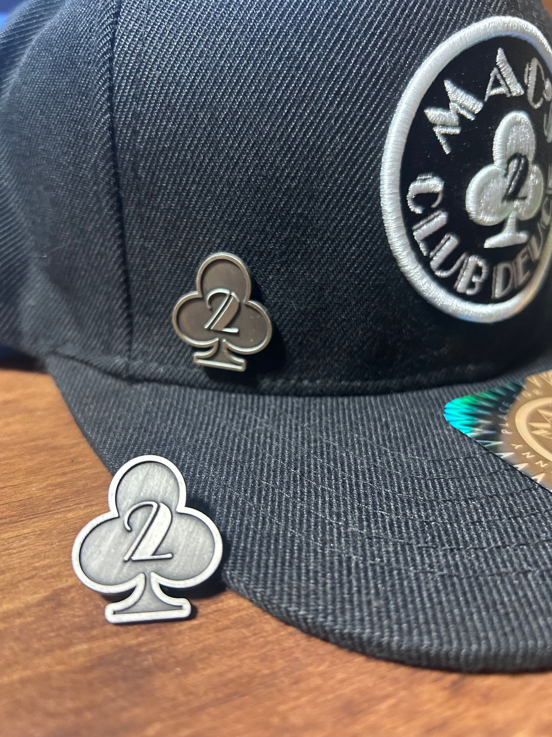Front of a small metal pin shaped as a club icon with the number two in the center. One on a hat and another beside the hat.