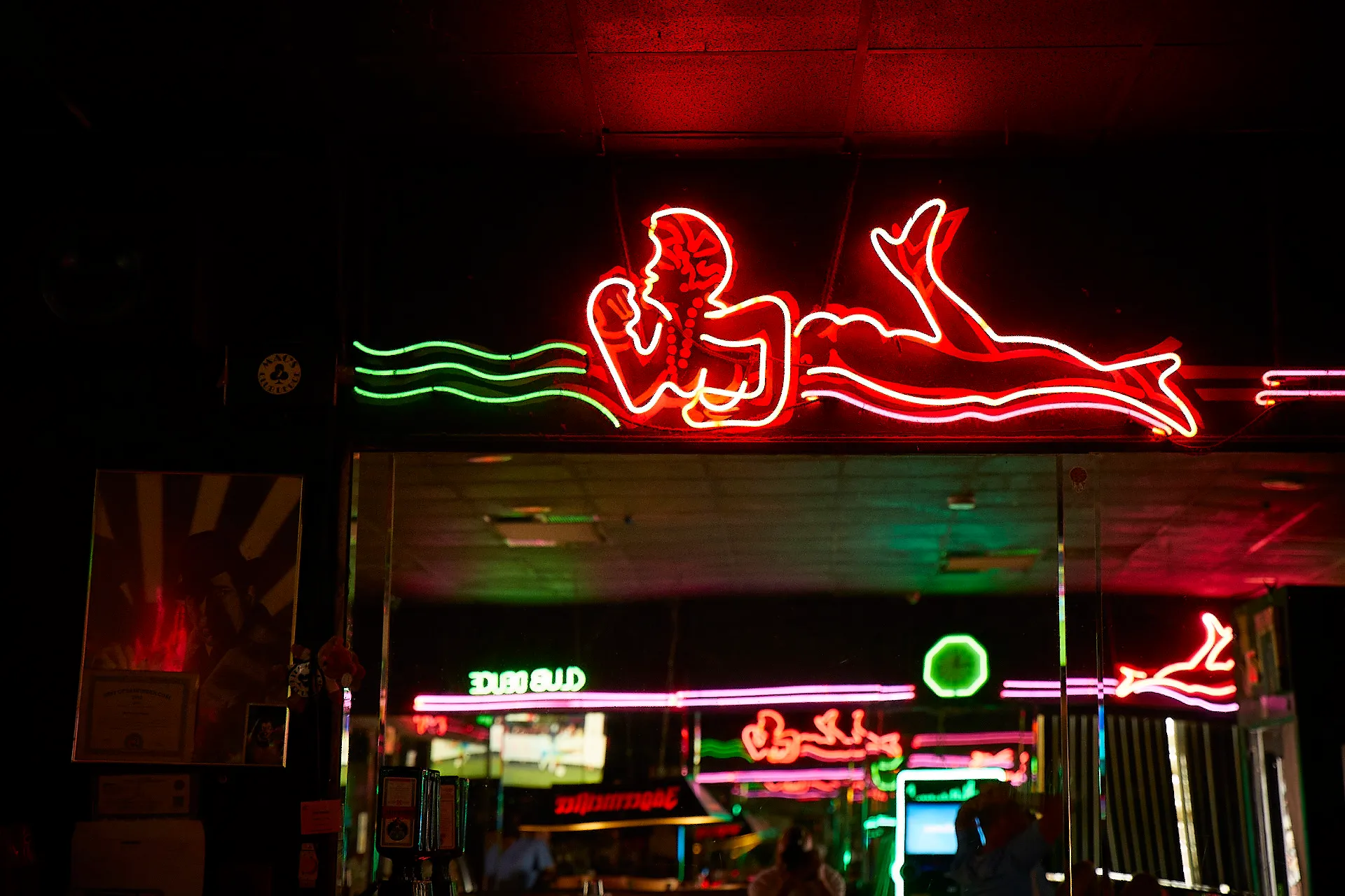  Mac's Club Deuce Beloved Neon Lady. The pink neon figure is surrounded by green and pink neon stripes. A mirror shows the reflection of another mirror from across the bar.