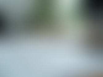 A close-up, shallow depth of field shot of a bright, minimalist wooden desk. In the foreground, a sheet of paper displays various hand-drawn wireframe sketches for app or website interfaces. In the blurred background, a silver laptop, a white keyboard, a smartphone, and a desktop computer monitor with an Apple logo are visible, suggesting a creative or design workspace.