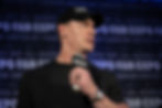 Man in black hat speaking into mic labeled "FANEXPO" against text-filled backdrop. Serious expression, black shirt, dim lighting.