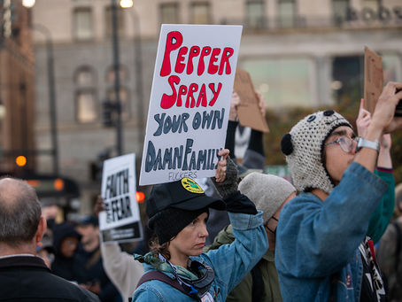 A photo of a person in a group of protesters holding a sign that reads "pepper spray your own damn family fuckers."