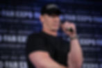 Man in a black shirt and cap holding a microphone on stage, with "FAN EXPO" text in background. Serious expression, dim lighting.