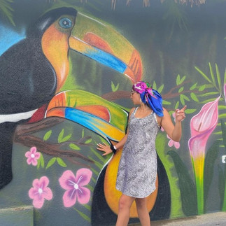 Girl in sundress with a headscarf posing in front of a mural of a couple of toucans and greenery.