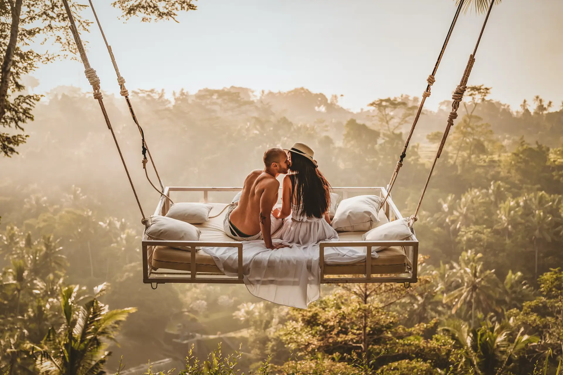 Couple sharing a kiss on a bed swing in Bali with lush greenery under them. 