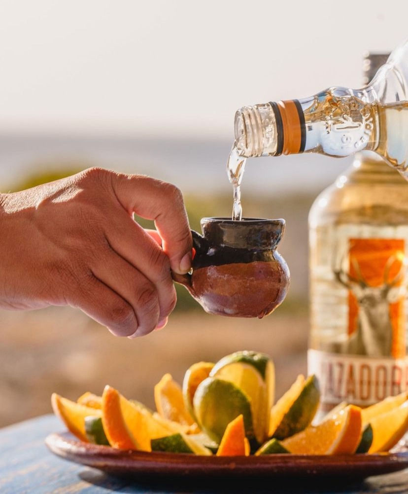 Mezcal being poured into a cute shot glass over a plate of orange slices.