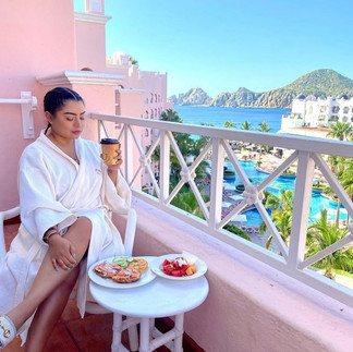 Woman in a white robe enjoying breakfast bagels and coffee on the balcony of her hotel room at Pueblo Bonito Rose, Medano Beach in Cabo Mexico in the background.