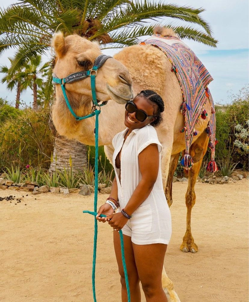 Girl posing with camel who's giving her a little kiss.