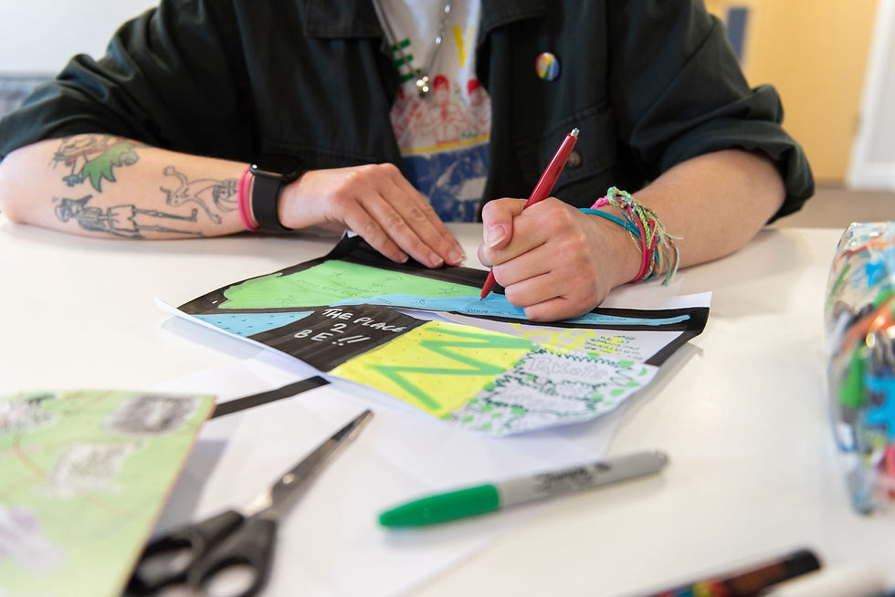 Image of the chest and arms of a white adult wearing a t-shirt and black top, with tattoos on one arm, drawing a picture and writing words on a table.