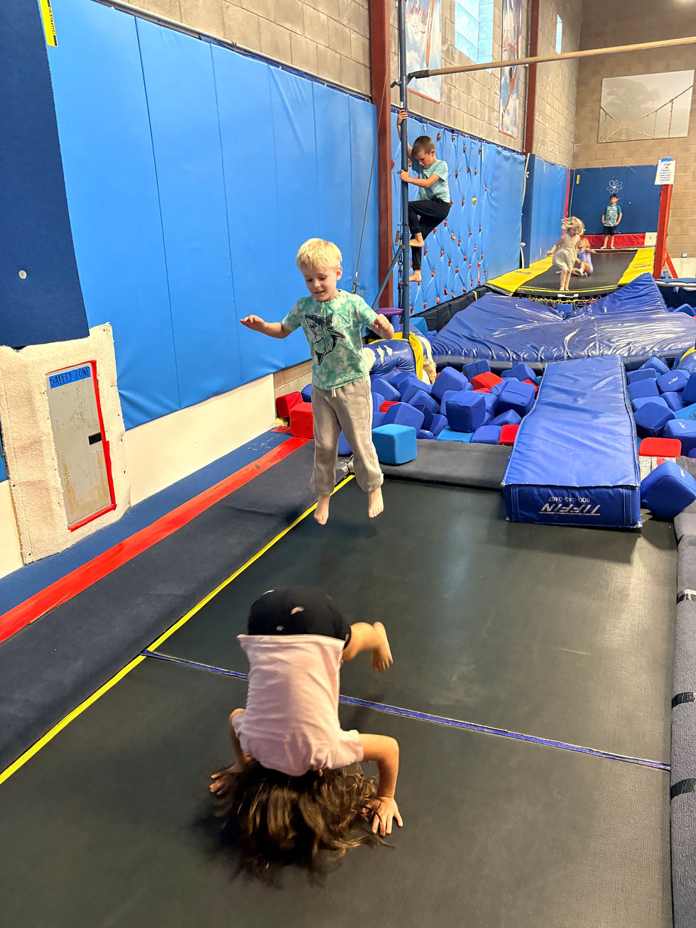 Kids play on trampolines and foam blocks in an indoor gym; one child balances, another does a headstand, with blue padded walls.