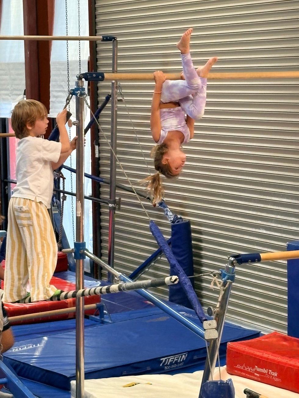 Child upside down on gymnastics bars while another child spots her in a gym.