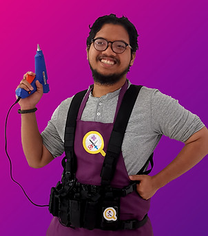 A half-body portrait of Yashi, a Malay male in his 20's. He has black facial hair and glasses. He is grinning widely, with a hot glue gun in his right hand, and his left hand on his hip. He is wearing a grey collared shirt underneath the Qiqi's Cosplay Medics purple apron and a tools harness.