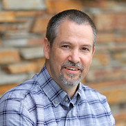 A headshot photo of Dr. John Whittaker smiling and wearing a blue plaid collared shirt.