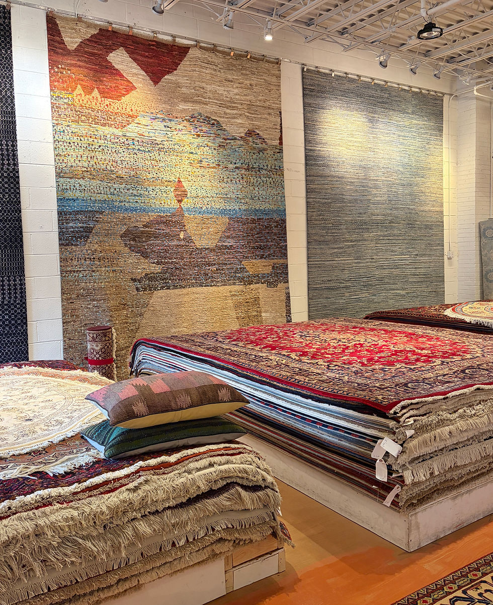 Rug showroom with stacked colorful patterned carpets and pillows under large hanging rugs on white walls and ceiling lights