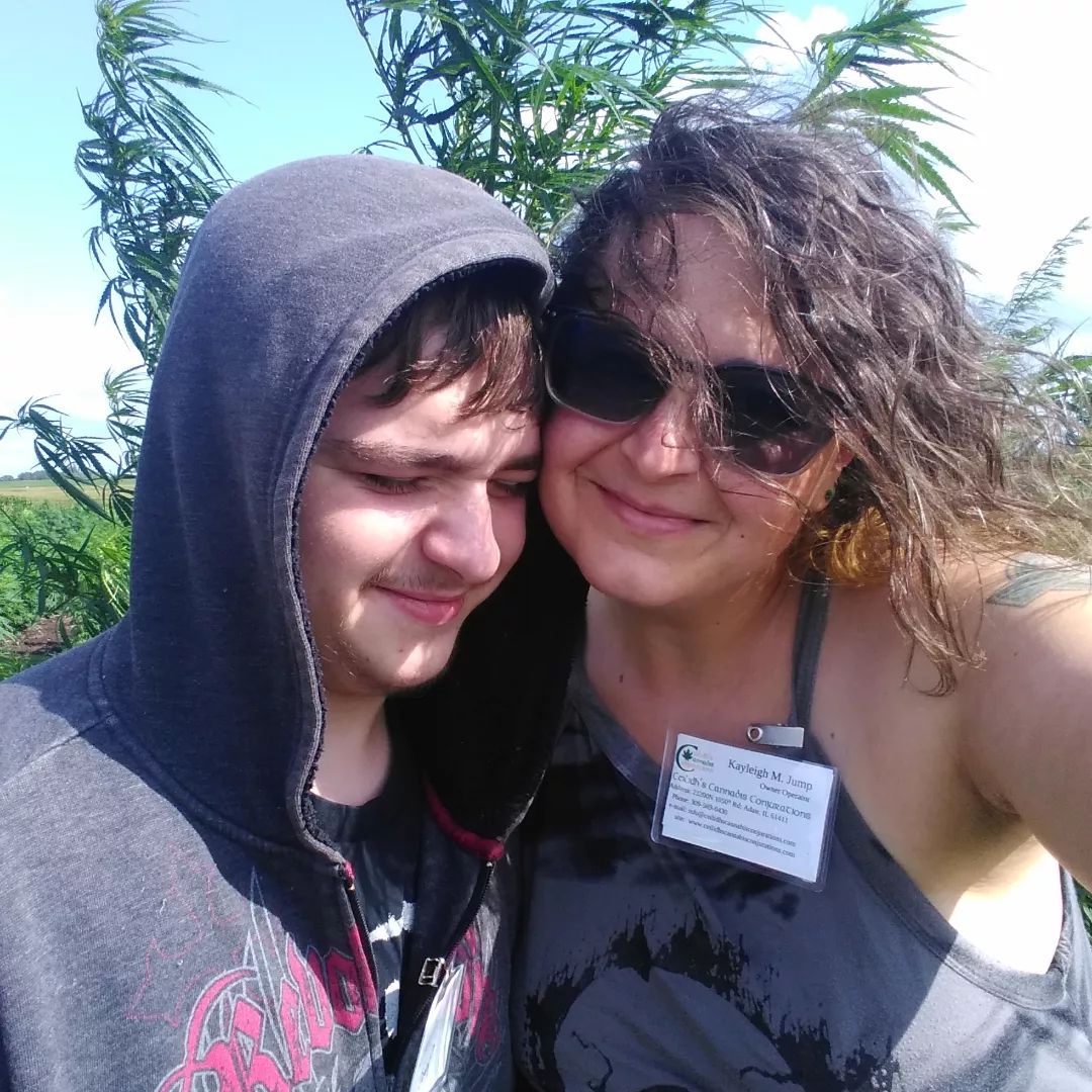 My son and I with feral hemp. Photo of woman in her late 30's with dark curly medium length hair, and a light olive skin tone.  She is wearing sunglasses and a grey tank top, a tattoo of a Celtic cross is partially visible on her left shoulder. She is looking at the camera. The son is teenaged. He has short medium brown hair and a peach fuzz mustache.  He is wearing a black shirt and grey hooded sweatshirt over his head.  He's turned into his mother with his eyes closed, the sun is shinning in his eyes.