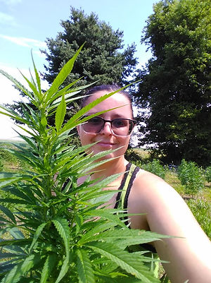 Me with dark hair pulled back standing next to a hemp plant in the field that towers my height.